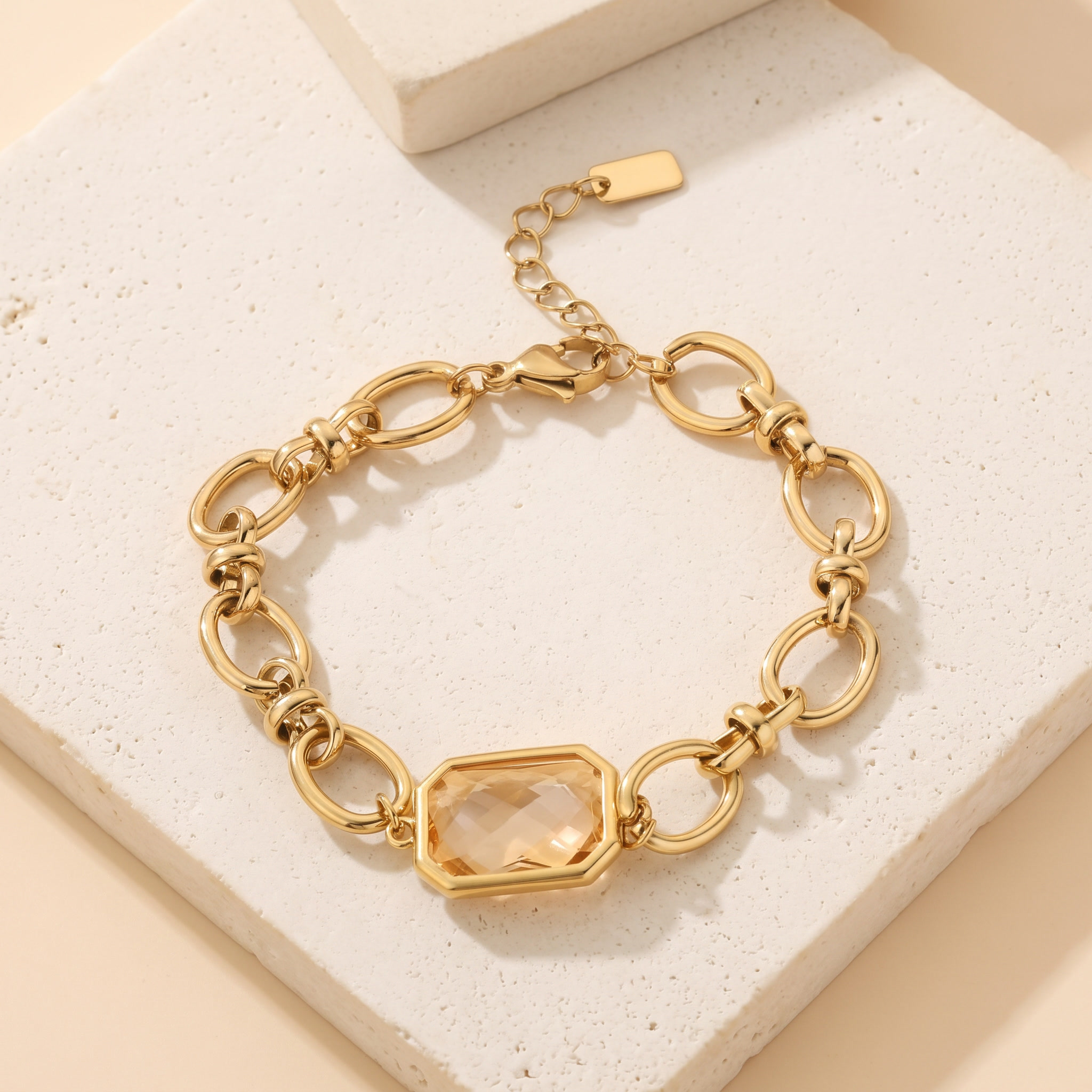 Bracelet Apogée en acier inoxydable doré, gros maillons et pierre octogonale champagne, packshot sur travertin ivoire, Maison Sveltesse