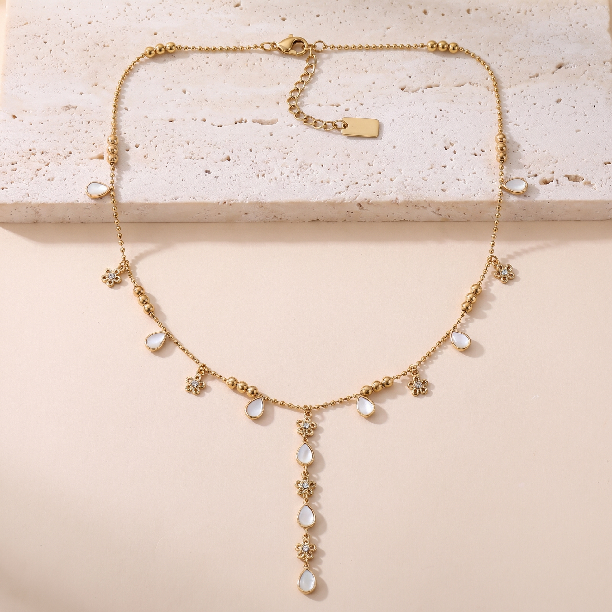 Collier Clair de Lune en acier inoxydable doré, chaîne à billes avec breloques nacre et fleurs, packshot sur travertin ivoire, Maison Sveltesse