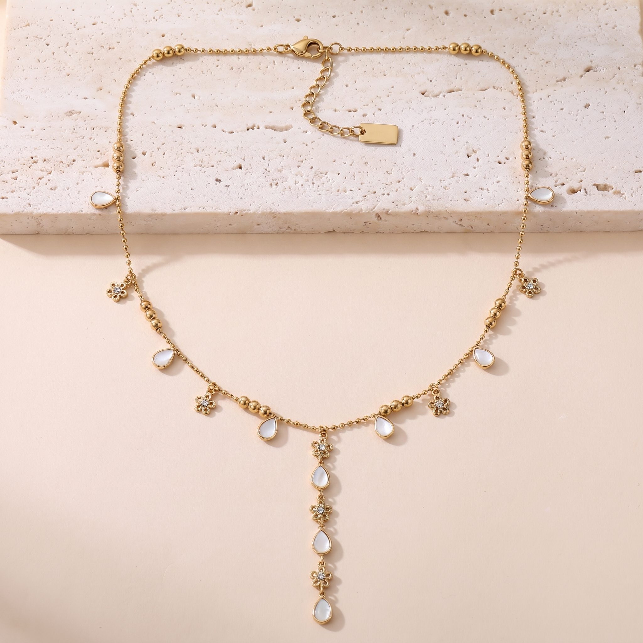 Collier Clair de Lune en acier inoxydable doré, chaîne à billes avec breloques nacre et fleurs, packshot sur travertin ivoire, Maison Sveltesse