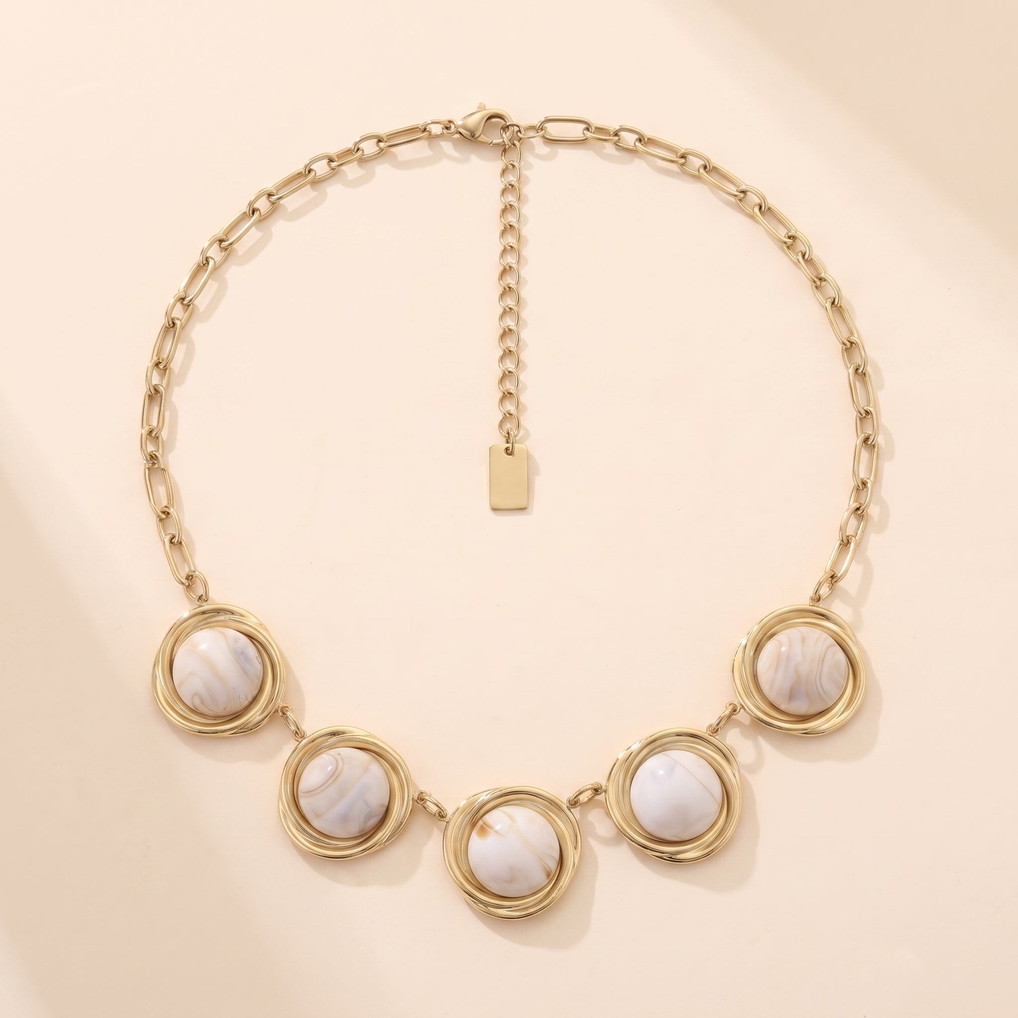 Collier Couchant Ivoire en acier inoxydable doré, cabochons marbrés crème et anneaux torsadés, packshot sur travertin ivoire, Maison Sveltesse