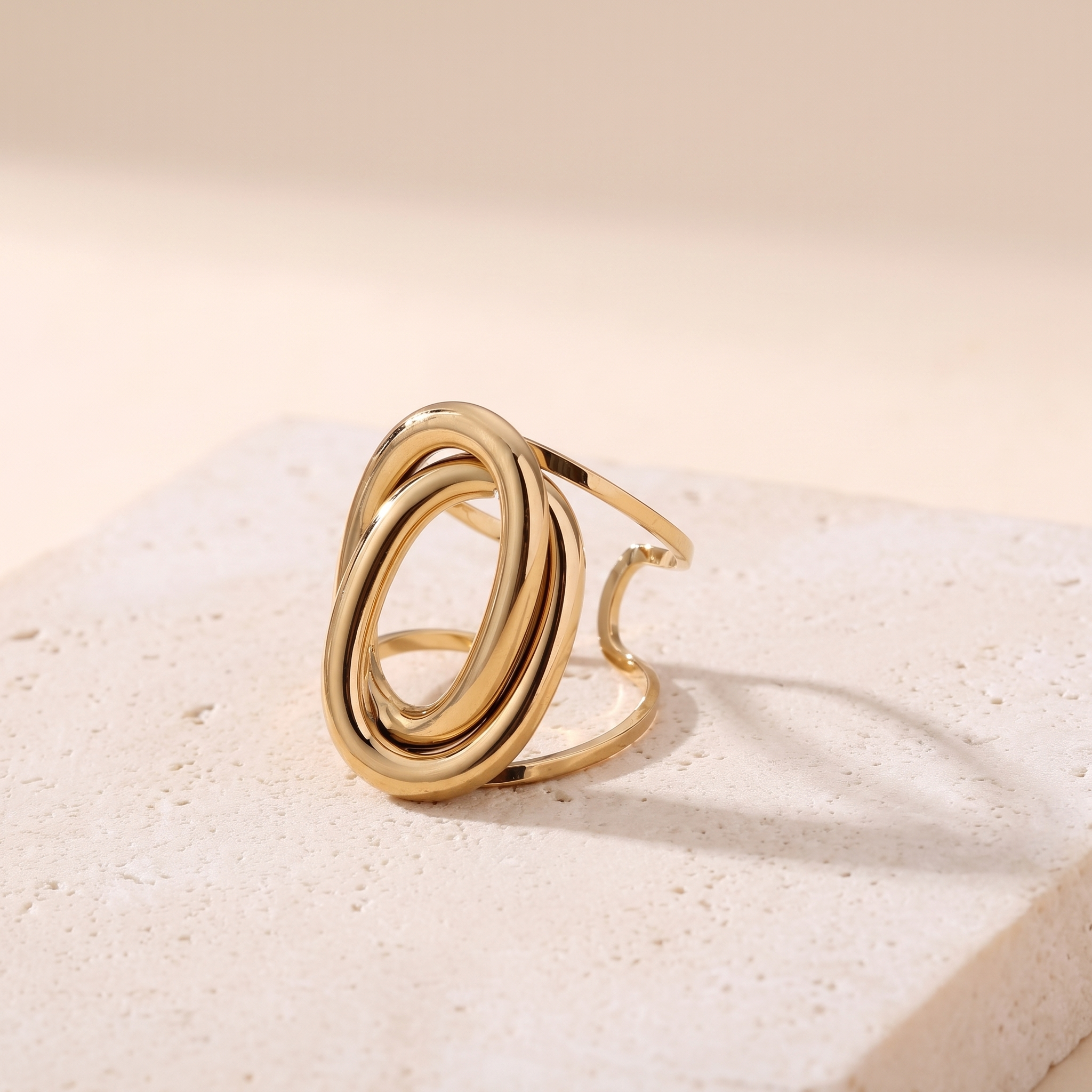 Bague Gémeaux en acier inoxydable doré, deux anneaux ovales entrelacés, packshot sur travertin ivoire, Maison Sveltesse
